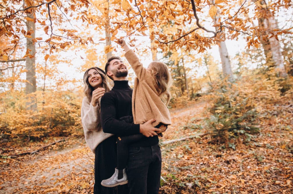 A family of three including a father, mother and young daughter in an autumn forest with golden fallen leaves, all looking upward and smiling at the tree canopy above, surrounded by trees with orange and yellow foliage.