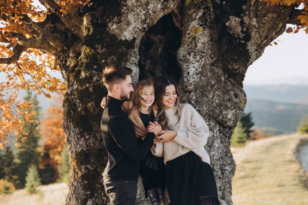 A young family of three, including a father, mother and daughter, standing together next to the trunk of a large old tree covered in moss and lichen, with autumn foliage and mountain landscape visible in the background.
