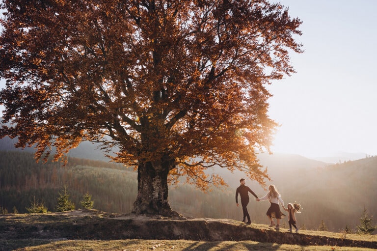 Silhouette of a family of three including a father, mother and young daughter, holding hands beneath a large tree with reddish-brown autumn foliage, backlit by golden sunlight in a misty field.