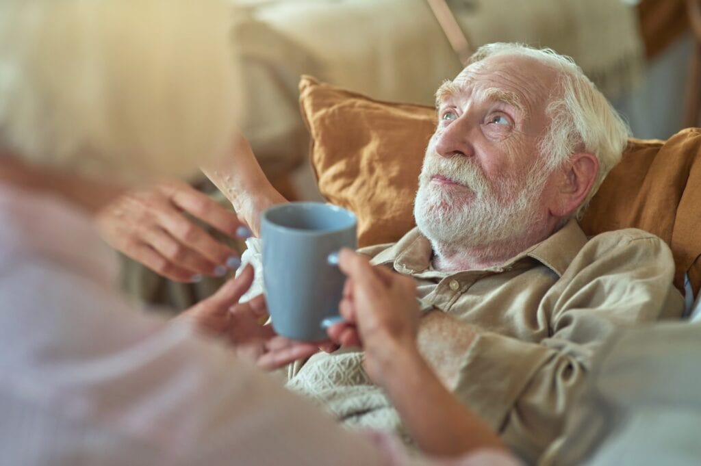 Woman giving cup of warm tea to sick elderly man