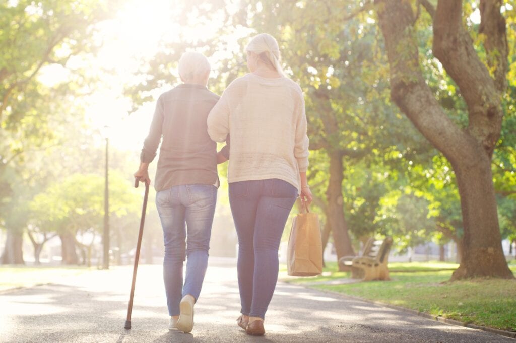A woman and her mother walk down a sunlit path with trees