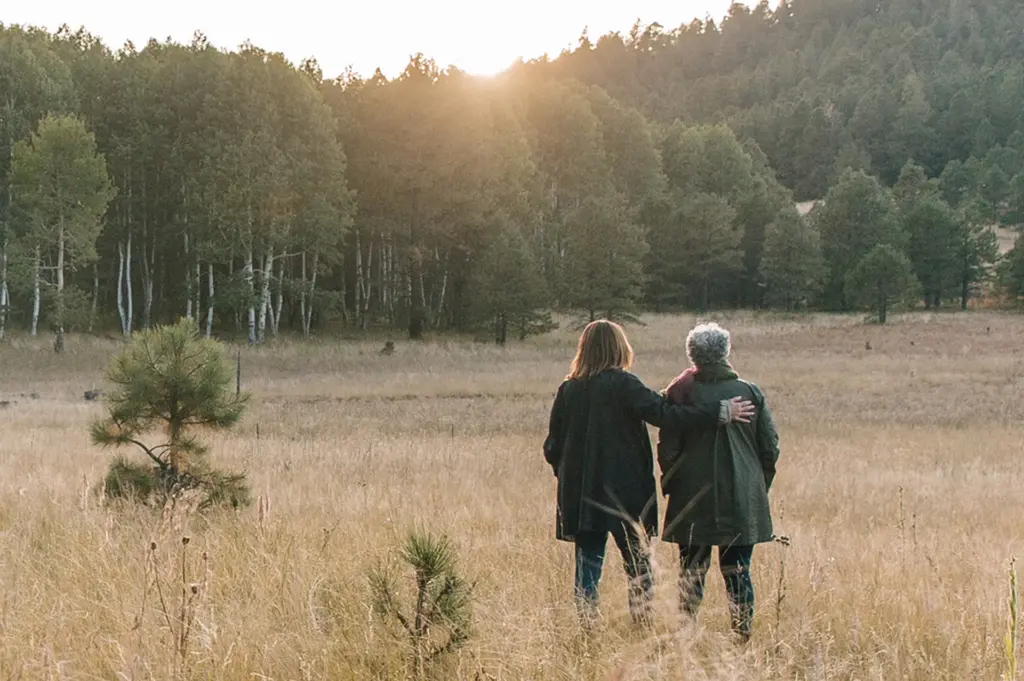 Back view of a senior mother and adult daughter  standing together gazing out at the forest, daughter's arm resting on mother's shoulder.