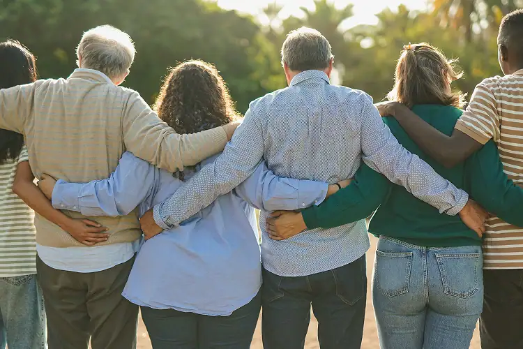 Back view of a diverse, multigenerational group hugging in a city park, symbolizing compassion, community, and Everis’s partnerships with Oakland hospice and healthcare organizations.