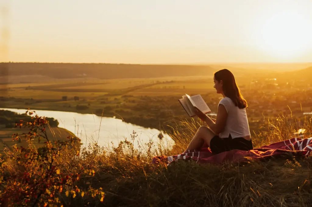 A woman reading a book at sunset in a meadow by a river
