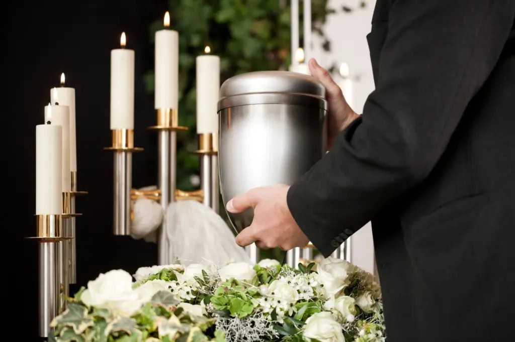 A hand reaches out to touch an urn displayed at a funeral surrounded by candles and flowers