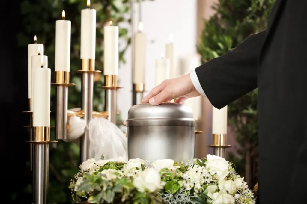 A hand reaches out to touch an urn displayed at a funeral surrounded by candles and flowers