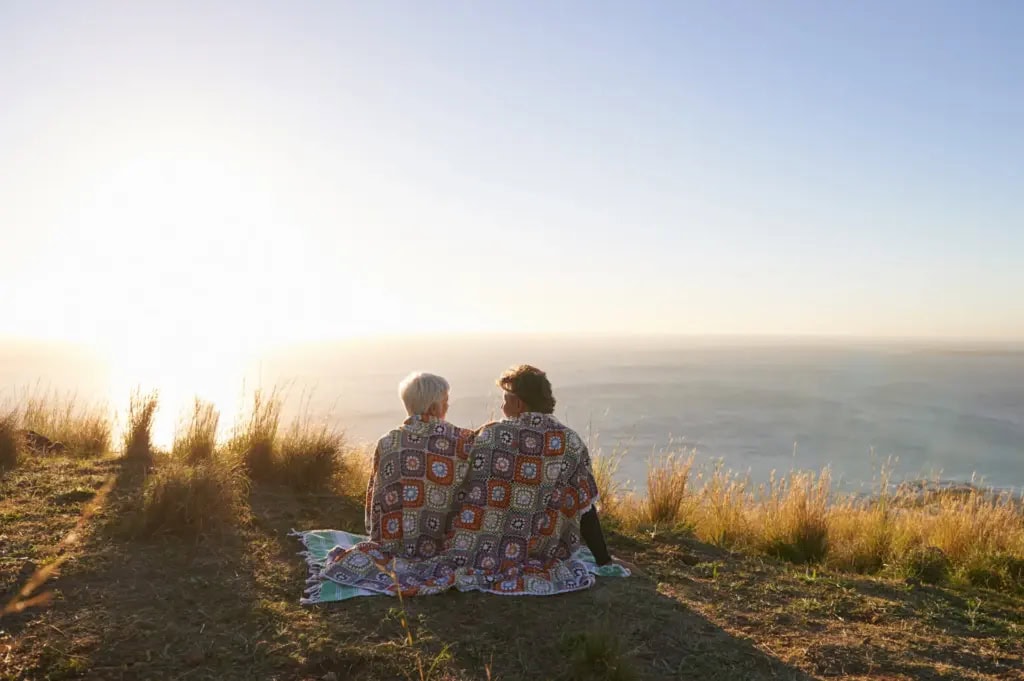Older couple sitting on a cliffside wrapped in a blanket looking at the sunset over the ocean