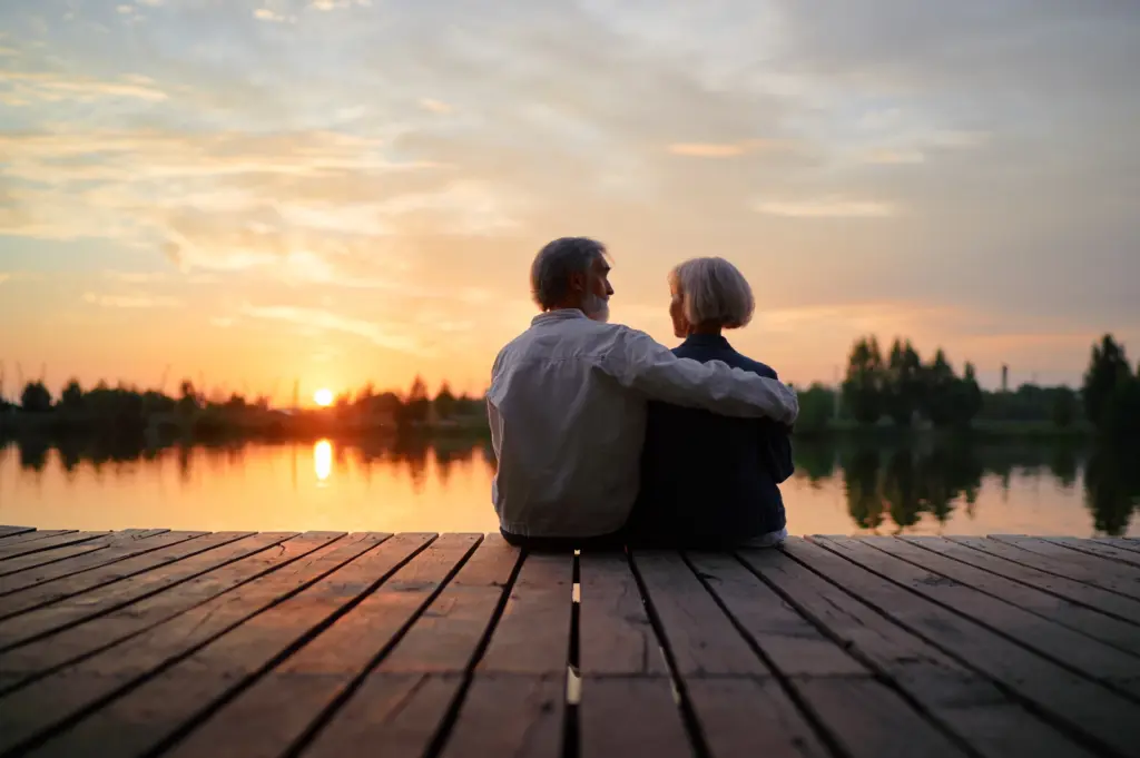 A Couple look at each other sitting on a dock with the sun rising over a lake