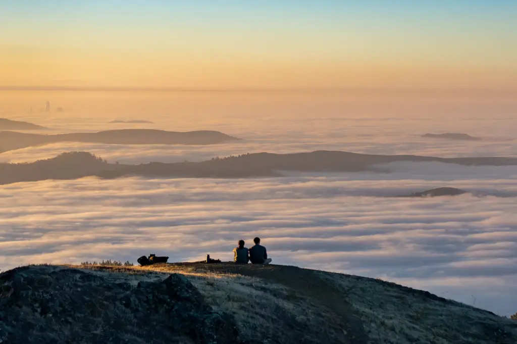 A couple sitting on hillside look out at clouds blanketing the earth below.