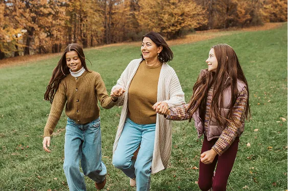 Grandmother walking between her two granddaughters, holding their hands as they walk down a grassy hill in a forest setting.