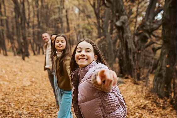 A family linked arm-in-arm smiling, with two young daughters in the foreground and mother and father in the background in a peaceful forest setting.