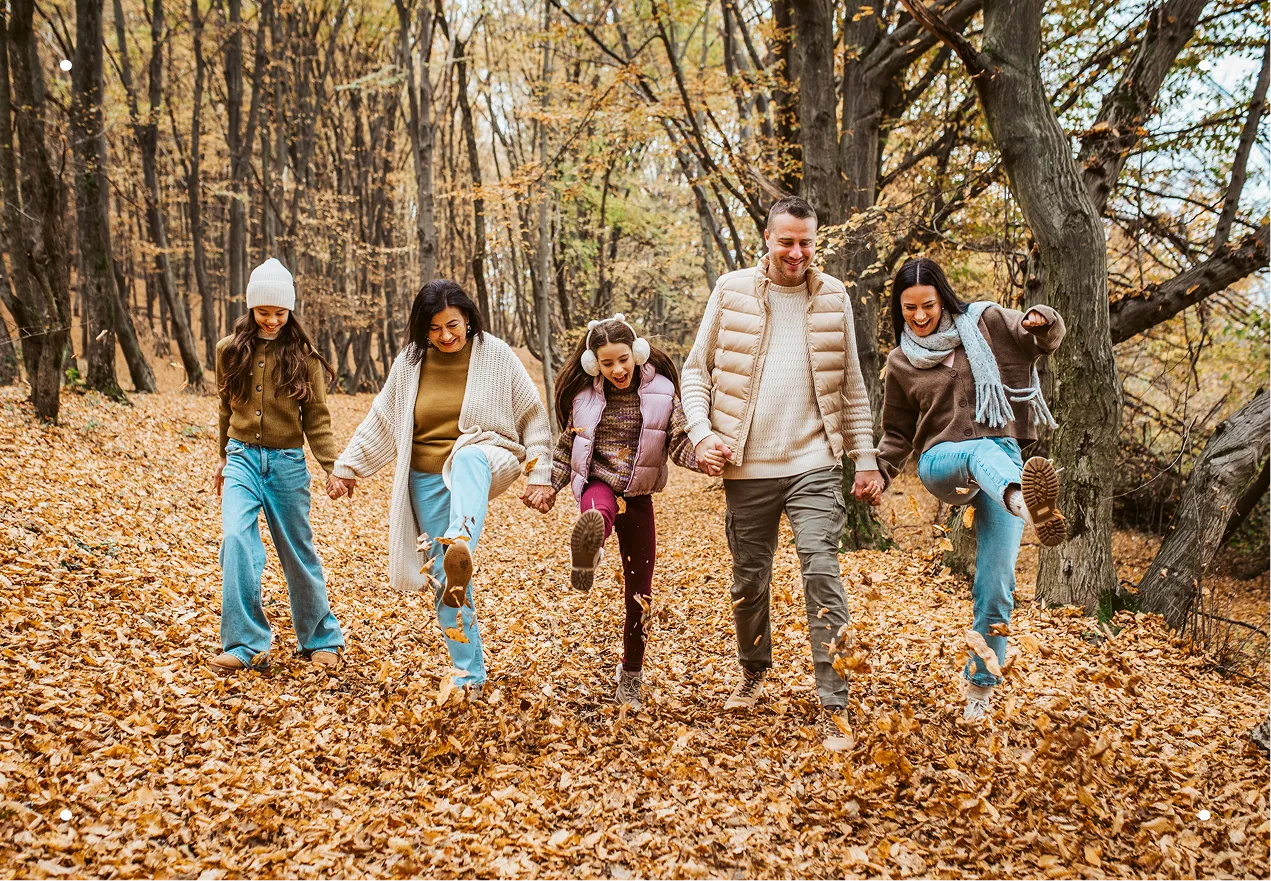 Multigenerational family including a grandmother, father, mother, and two young daughters linked arm-in-arm, smiling as they playfully kick up leaves as they walk through a peaceful forest setting on a cold winter day.