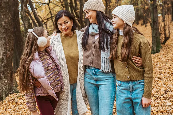 Multigenerational family including a grandmother, mother, and two young daughters linked arm-in-arm, smiling at each other in a peaceful forest setting on a cold winter day.