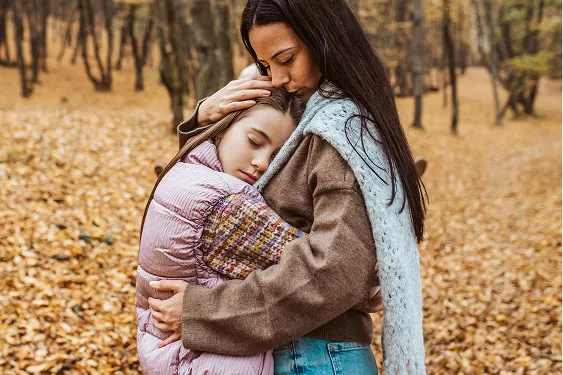 Mother and young daughter sharing a tender hug with their eyes closed in a peaceful forest setting.