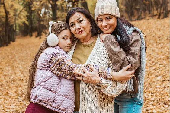 Multigenerational family including a grandmother, mother, and a young daughter, smile and embrace each other in a peaceful forest setting on a cold winter day.