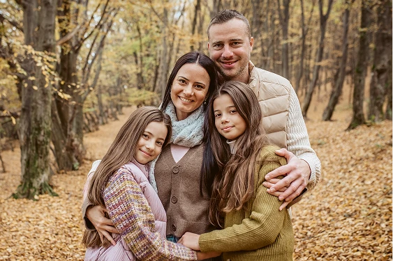 Portrait of mother, father and two tween daughters embracing in the foreground with the forest in the background.