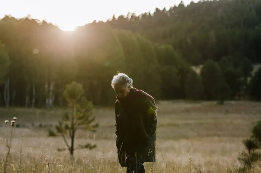 Senior woman walks through a peaceful field with forest in the background at sunset, looking down as if deep in thought.