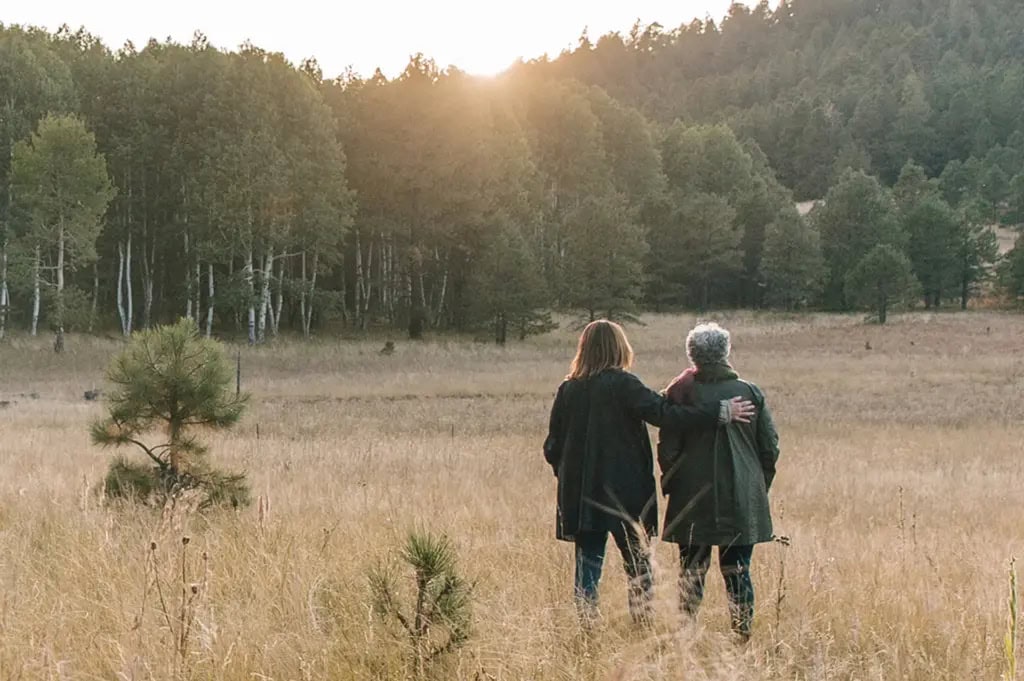 Back view of a senior mother and adult daughter standing together gazing out at the forest, daughter's arm resting on mother's shoulder.