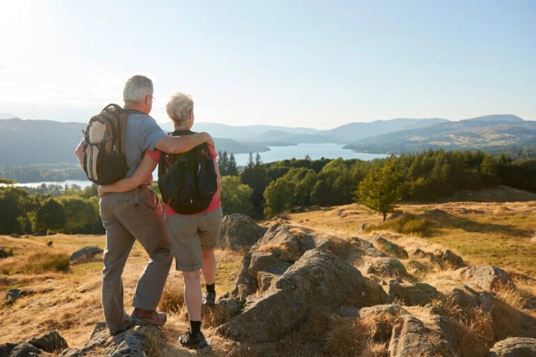 An older couple standing on a rocky hillside overlooking a scenic lake and mountains, with golden grass and autumn-colored landscape stretching into the distance.