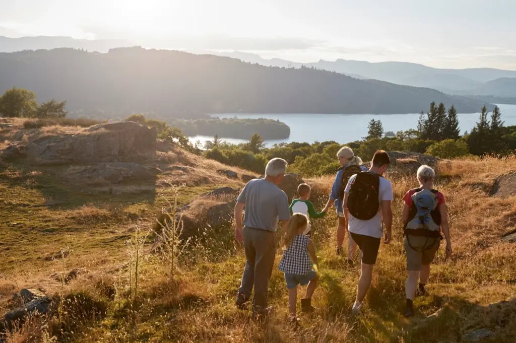 Back view of a multigenerational family including grandparents, parents, and a young son and daughter hiking through a dry grassy hillside toward a body of water in the distance.