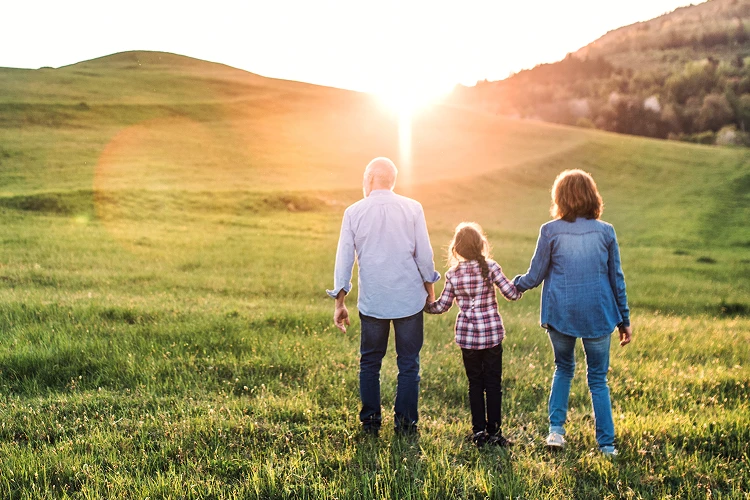 A grandfather, mother, and daughter walk hand in hand across a grassy field towards a beautiful setting sun.
