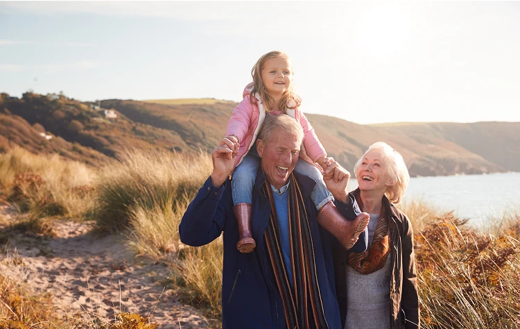 A senior couple walk side by side with their granddaughter sitting on the grandfather's shoulders, with a scenic landscape of rolling hills and a body of water behind them.