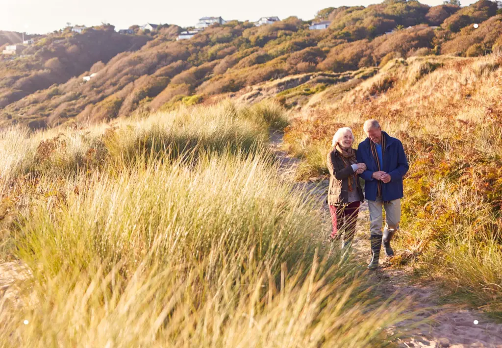 A senior couple walking arm-in-arm down a sunny hillside path surrounded by tall dry grass.