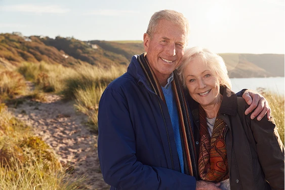A happy senior couple in a warm embrace, smiling, with a scenic landscape of rolling hills and a body of water behind them.