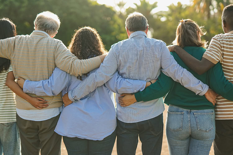 Diverse group of people (various ages and ethnicities) embracing each other in an outdoor setting.