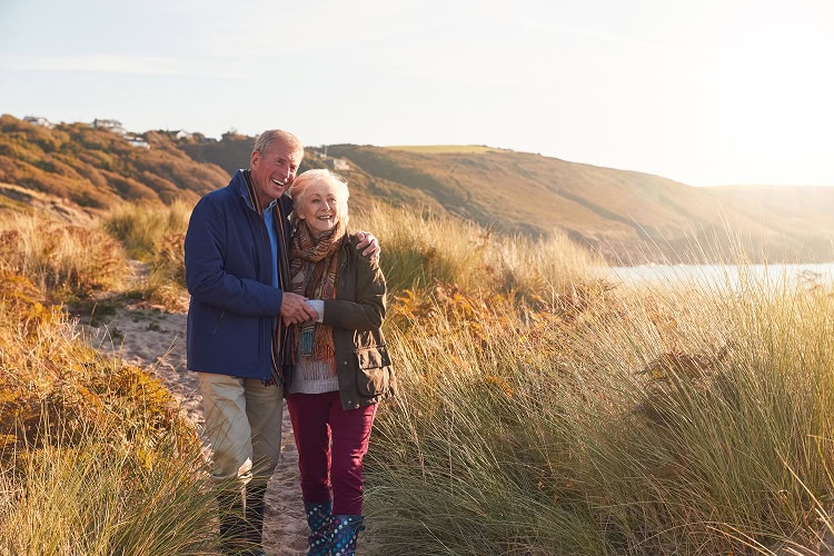 A senior couple walking arm-in-arm down a sunny hillside path surrounded by tall dry grass, pausing to observe something in the distance.