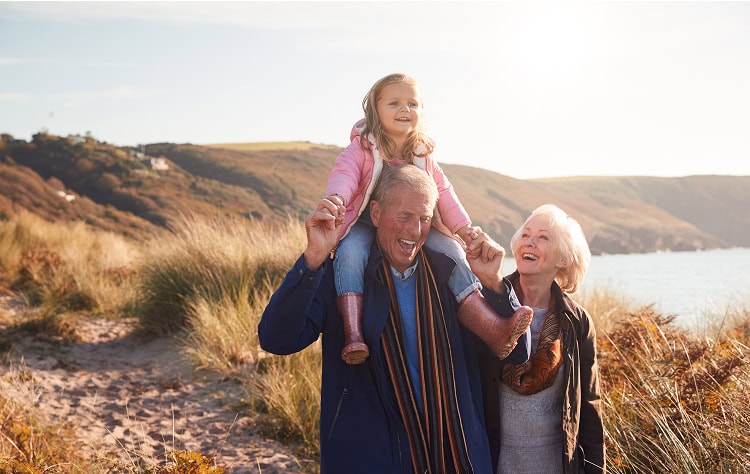 A senior couple walk side by side with their granddaughter sitting on the grandfather's shoulders, with a scenic landscape of rolling hills and a body of water behind them.