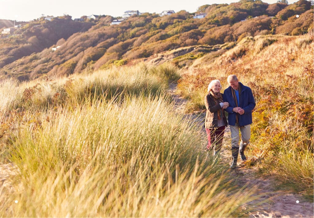 A senior couple walking arm-in-arm down a sunny hillside path surrounded by tall dry grass.
