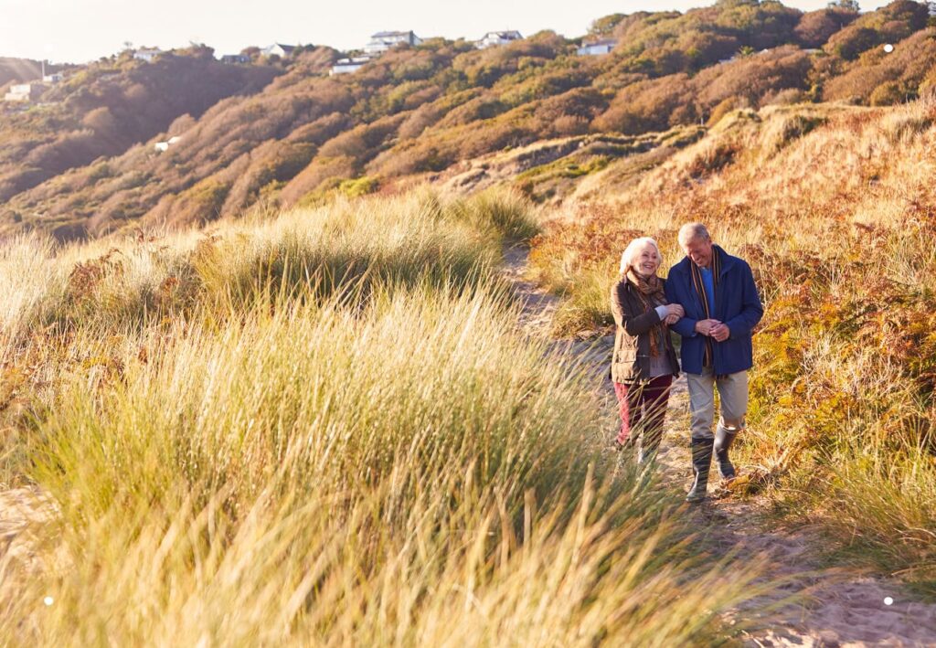 A senior couple walking arm-in-arm down a sunny hillside path surrounded by tall dry grass.