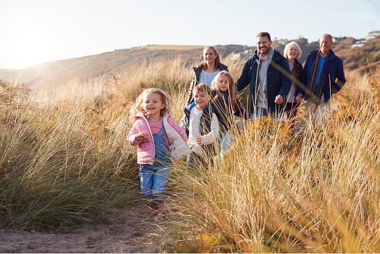 Happy multigenerational family of children, parents, and grandparents running together down a sunny hillside covered in tall dry grass, with three small children leading the way.