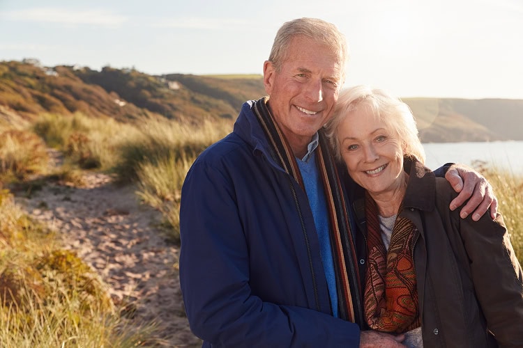 A happy senior couple in a warm embrace, smiling, with a scenic landscape of rolling hills and a body of water behind them.