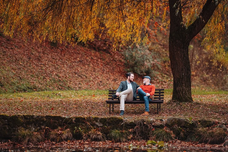 Adult son and older father sitting on a bench by a peaceful lake, talking and finding comfort together.