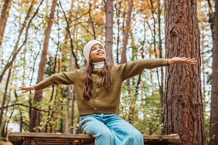 Tween girl sitting on a bench in the forest with her arms stretched out, feeling the breeze on a cool winter day.