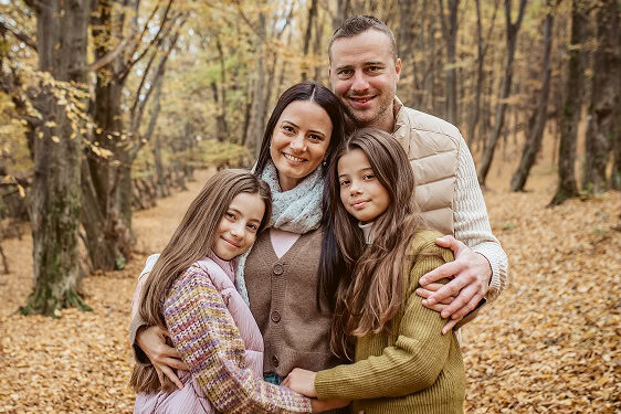 Portrait of mother, father and two tween daughters embracing in the foreground with the forest in the background.