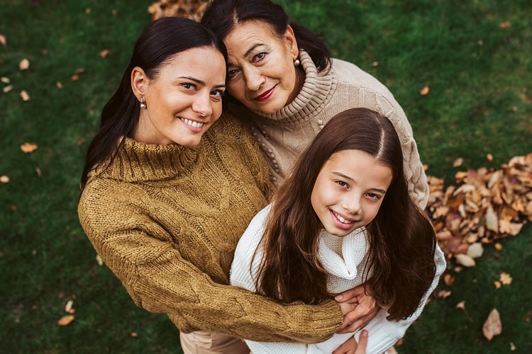 Portrait from above of a grandmother, mother, and daughter embracing and smiling in an outdoor setting.