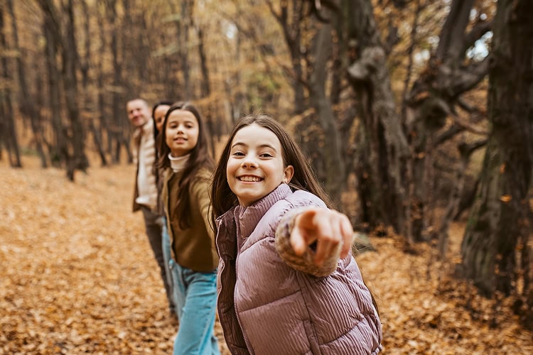 A family linked arm-in-arm smiling, with two young daughters in the foreground and mother and father in the background in a peaceful forest setting.