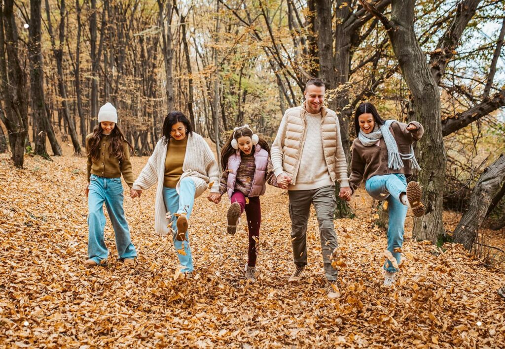 Multigenerational family including a grandmother, father, mother, and two young daughters linked arm-in-arm, smiling as they playfully kick up leaves as they walk through a peaceful forest setting on a cold winter day.