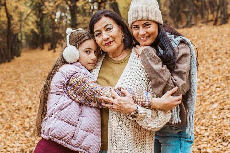 Multigenerational family including a grandmother, mother, and a young daughter, smile and embrace each other in a peaceful forest setting on a cold winter day.
