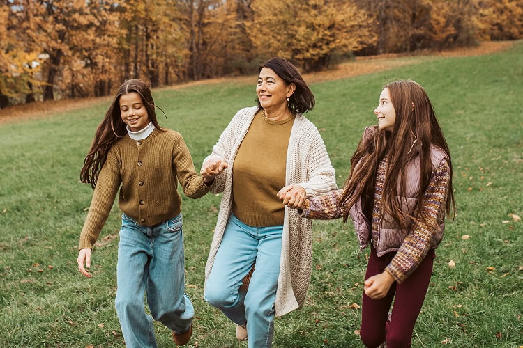 Grandmother walking between her two granddaughters, holding their hands as they walk down a grassy hill in a forest setting.