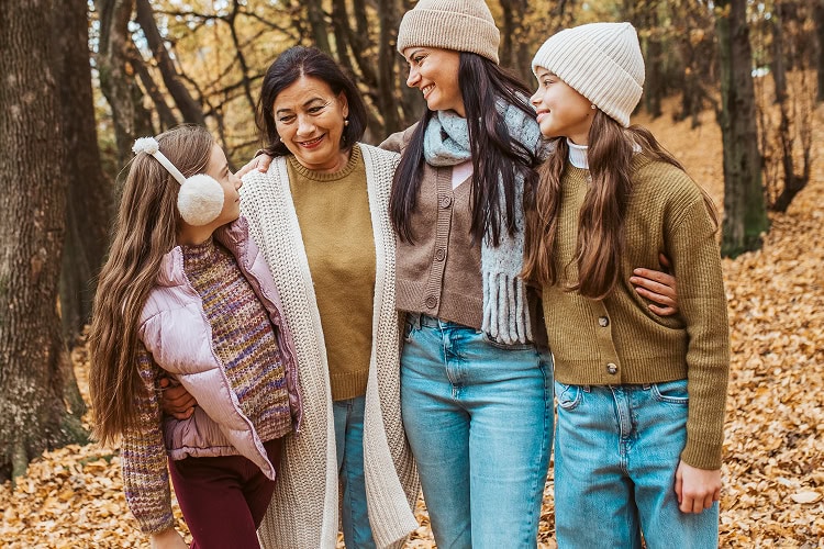 Multigenerational family including a grandmother, mother, and two young daughters linked arm-in-arm, smiling at each other in a peaceful forest setting on a cold winter day.