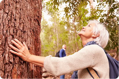 An older woman with white hair resting her hand on a tree trunk while looking upward with a delighted expression at the Memorial Tree.