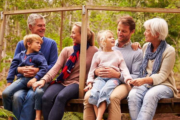 A multi-generational family sitting on wooden bridge with legs hanging over the edge, enjoying time together in forest setting.