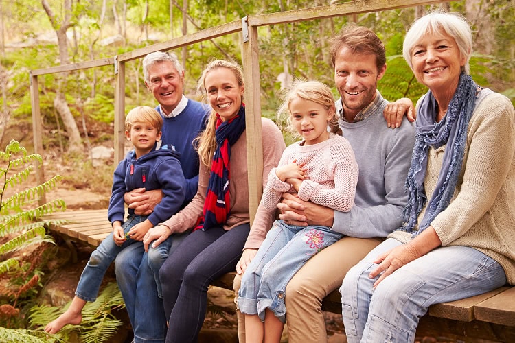 A multi-generational family sitting on wooden bridge with legs hanging over the edge, enjoying time together in forest setting.
