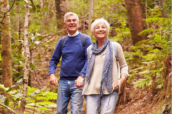 An older couple, each with white hair, walk hand in hand through the forest.