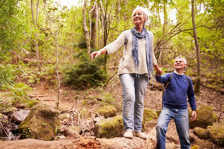 Smiling older woman balances on a fallen tree trunk while her partner holds her hand on a forest trail.