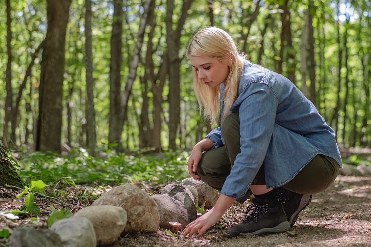 Woman kneeling beside a rock-lined path with her hand near a memorial marker in a Spreading Grove, symbolizing remembrance and renewal in nature.